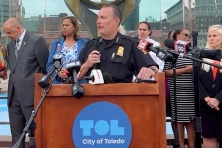 Toledo Police Chief speaks at a podium with microphones, flanked by diverse city officials and community members outdoors.