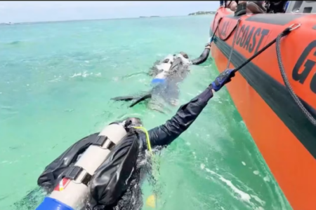 Two scuba divers in clear turquoise water next to a US Coast Guard boat, one holding a line, the other partially submerged.
