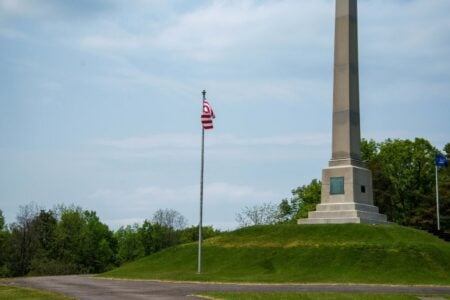 US flag and New York State flag flying near a tall obelisk monument on a grassy hill.