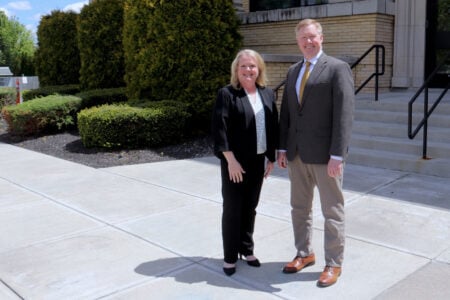 A smiling woman and man stand on a sidewalk outside a light brick building with steps and green hedges.