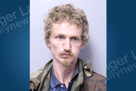 Blond man with curly hair, mustache, and goatee, wearing a blue shirt and camouflage jacket, in a police mugshot.