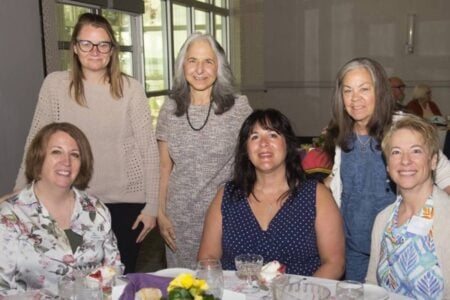 Six smiling women pose for a group photo at a table during an indoor event.