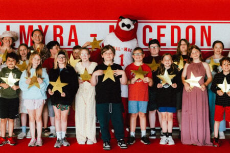 Palmyra-Macedon students and adults, with their mascot, hold golden star awards on a red carpet in front of their school banner.