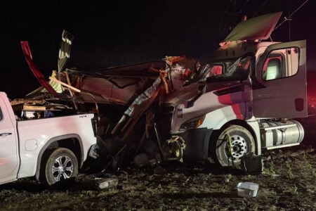 Severely damaged white pickup and semi-truck amidst collapsed building debris at night in a muddy field.