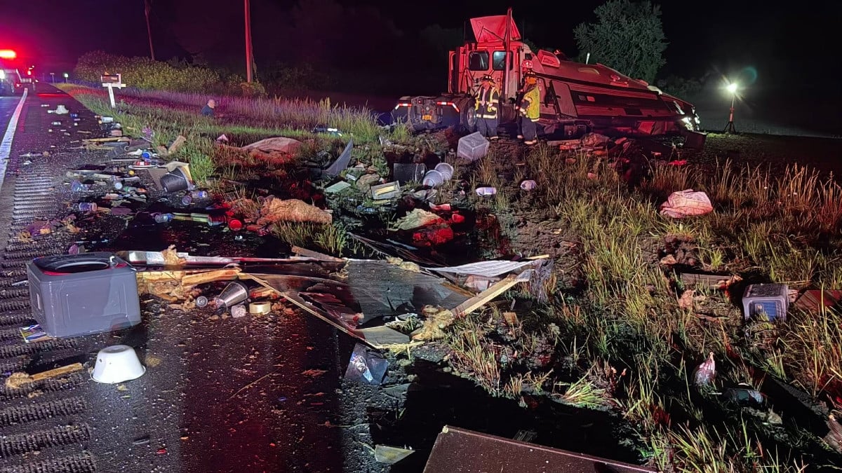 Wrecked truck cab and demolished trailer with debris littering a dark roadside field, attended by emergency workers.