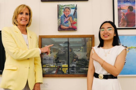 A smiling woman in a yellow blazer and a young woman in white point at a painting in a gallery.