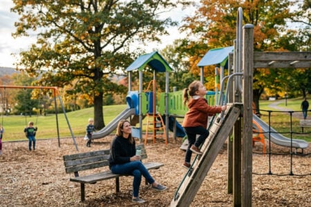 Woman on a bench watches a girl climb a playset while other children swing in an autumn park by a lake.
