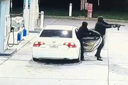 Two individuals at a gas station: one aims a rifle from a white sedan, while another partially exits the car.
