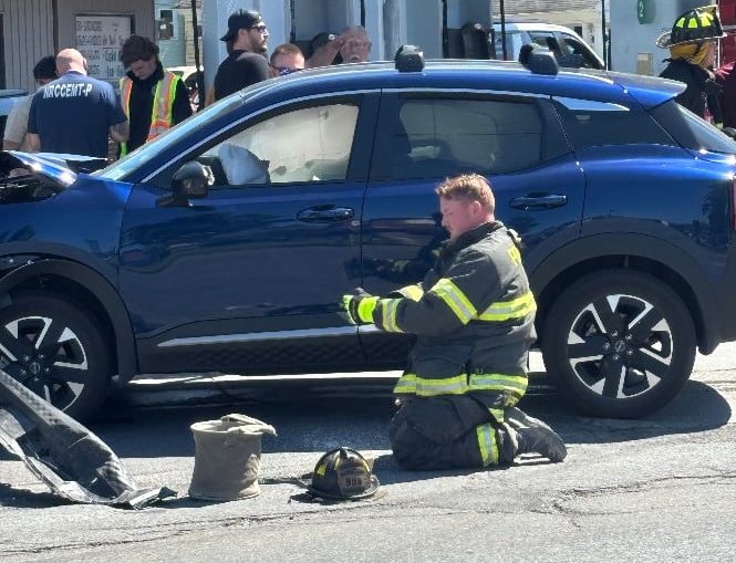 A firefighter kneels, working on a damaged blue car with deployed airbags at an accident scene.