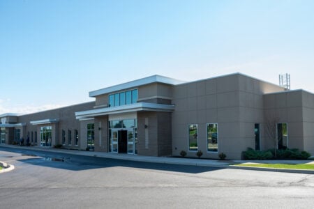 Modern beige office building with brick accents, glass entrances, and a paved parking lot on a sunny day.