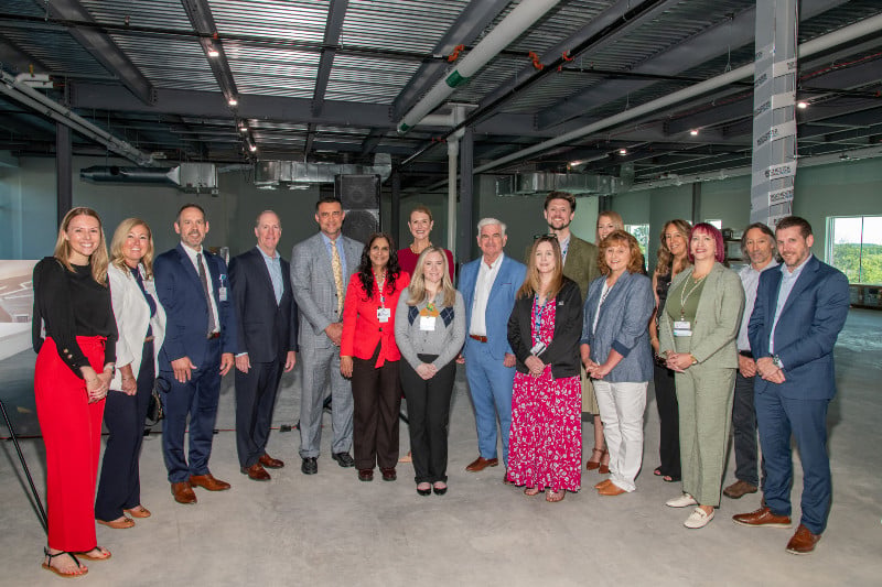 Nineteen people stand together in a large, newly constructed space with an exposed ceiling.