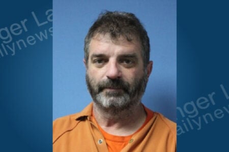 Mugshot of a man with a gray beard and dark hair wearing an orange uniform against a blue background.