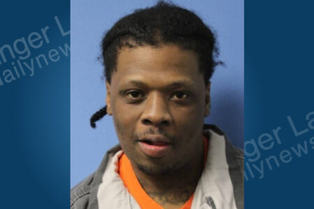 Man with dark skin, short braided hair, and facial hair, wearing an orange and grey shirt, looks forward in a mugshot.