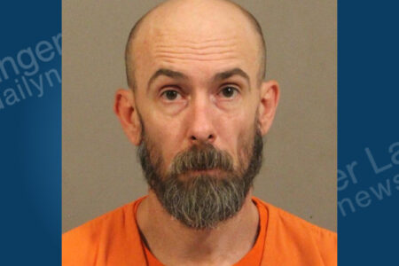 Mugshot of a balding man with a dark beard, wearing an orange shirt, looking directly at the camera.