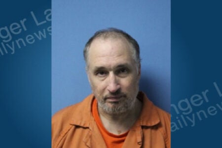 Mugshot of a middle-aged man with a balding head and beard, wearing an orange jumpsuit against a blue background.