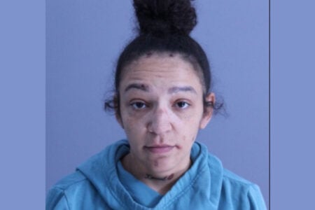 A woman with dark hair in a bun, blue hoodie, neck tattoo, and facial marks, looks directly at the camera on a plain background.
