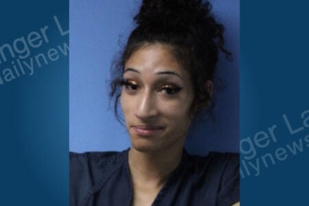 Person with dark curly hair in a bun, makeup, and a dark shirt, in a booking photo against a plain blue background.