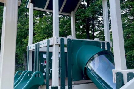 A new green and white playground structure with a clear tube slide and two open slides, surrounded by lush trees.