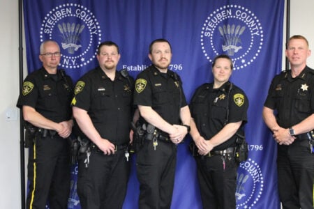 Five Steuben County Sheriff's deputies stand in uniform in front of a blue banner with the county seal.