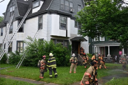 Syracuse firefighters set ladders against a large white house and connect hoses, with water spraying from a hydrant.