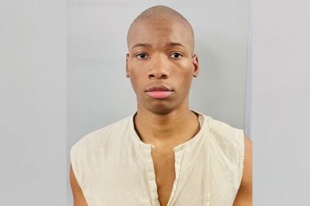 Young Black man with a shaved head, wearing a light beige sleeveless shirt, looking directly at the camera against a gray background.
