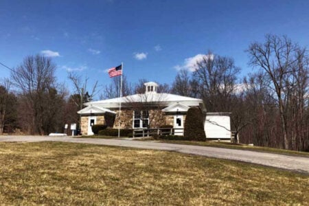 Stone building with a white roof, cupola, and US flag flying, set against bare trees and a blue sky.