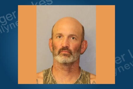 Bald man with a graying beard wearing a camouflage tank top in a mugshot against a light background.