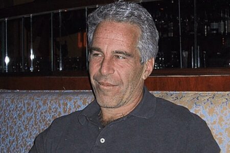 Jeffrey Epstein smiles slightly, seated on a patterned banquette in an indoor setting with a dark, reflective background.