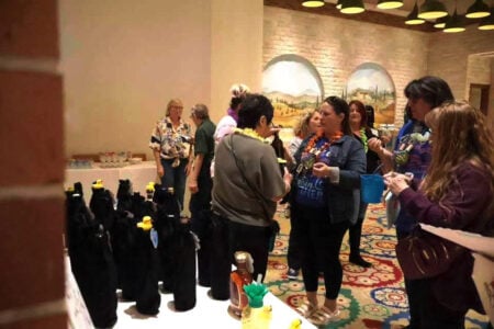 Guests wearing leis mingle at an indoor event with tables of black bottles and yellow ducks, under murals of arched landscapes.
