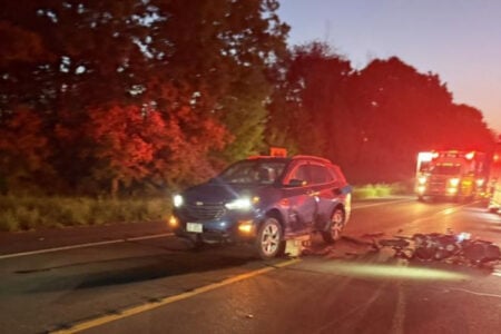 A dark blue SUV and a crashed motorcycle on a road at dusk, with emergency lights and an ambulance in the background.
