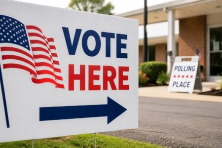 A white "VOTE HERE" sign with an American flag graphic and arrow on a lawn, leading to a polling place building.