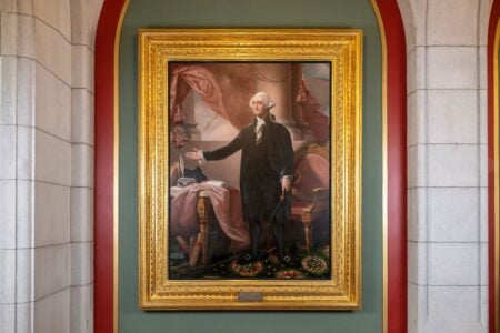 Ornate framed portrait of George Washington standing, displayed on a green wall between stone columns.