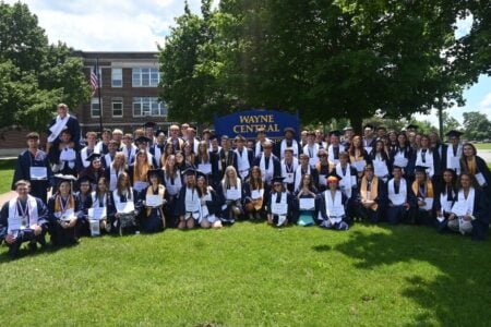 Wayne Central graduates in caps and gowns hold diplomas and pose for a group photo in front of their school.