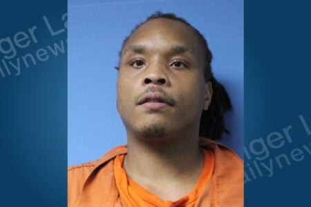 Man with dreadlocks and facial hair in an orange jumpsuit looks forward against a plain blue background.