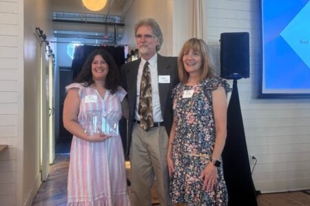 Three smiling PRSA members pose with a PRISM award at an indoor event, a screen behind them reads "Finger Lakes Country".