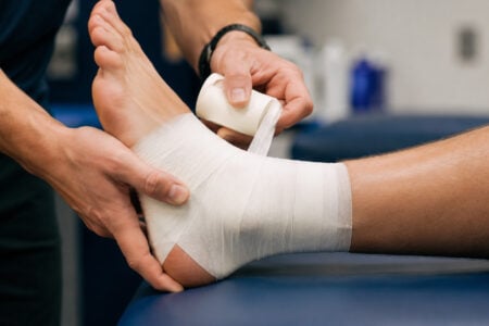 Hands wrapping a white bandage around a person's ankle on a blue examination table.