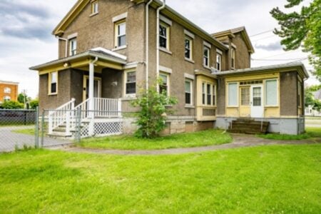 Old, stucco two-story house with yellow trim, a porch, and sunroom on a grassy lawn under a cloudy sky.