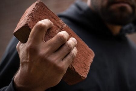 A person in a dark hoodie firmly grips a textured red brick, their bearded face blurred behind.