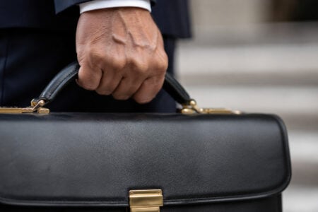 Close-up of a hand in a dark suit holding a black leather briefcase with gold clasps, blurred steps in background.