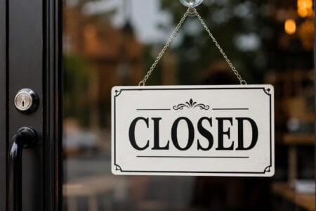 White "CLOSED" sign with black text hangs on a locked glass store door, reflecting blurred interior and exterior.