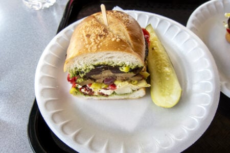 Vegetable pesto sandwich on an Italian roll with a pickle, served on a foam plate.