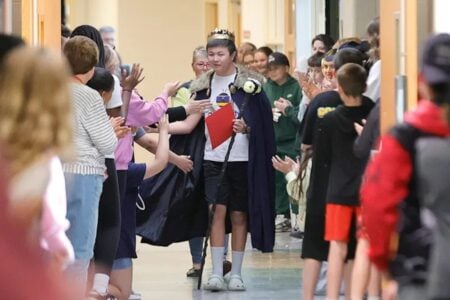 Student in a crown and cape walks down a school hallway past applauding classmates and teachers.