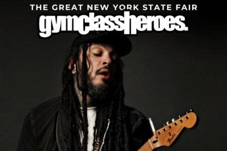 Gym Class Heroes frontman Travis McCoy sings with eyes closed, holding an electric guitar, against a dark background with fair branding.