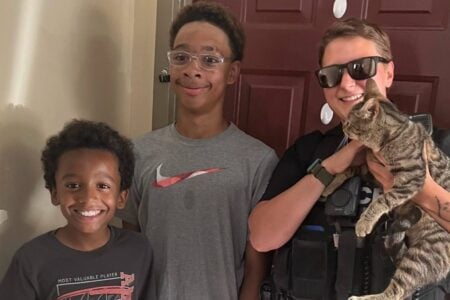 Smiling Seneca Falls officer holds a tabby cat with two happy boys at a home doorway.