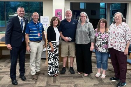 Seven adults, three men and four women, stand together and smile indoors at a library or community center.