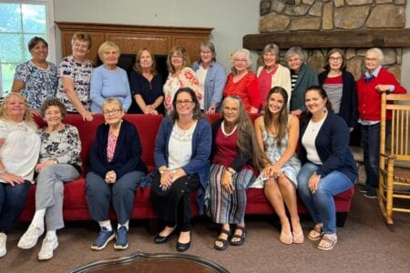 Twenty women of varying ages pose in a room with a stone fireplace; some sit on a red couch, others stand.