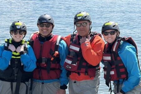 Four smiling people in water rescue gear, helmets, and life vests stand by the Finger Lakes.