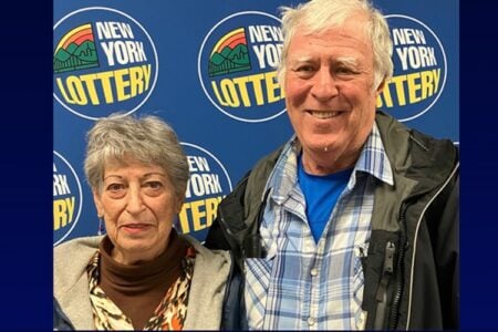 An older man smiles next to a woman, both standing in front of a blue New York Lottery logo backdrop.