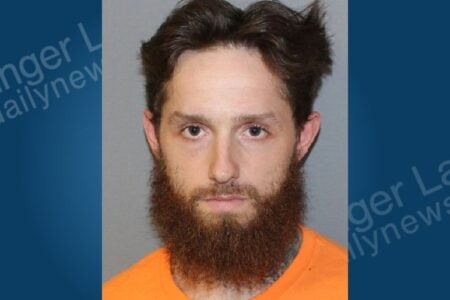 Mugshot of a man with brown hair, full beard, and an orange shirt, looking forward against a plain gray background.