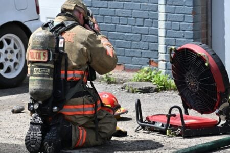 Firefighter in bunker gear and SCBA kneels on pavement next to a helmet and large red ventilation fan.
